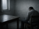 Gri ve karanlık bir odada, sırtı dönük şekilde sandalyede oturan yalnız bir adam; masanın üzerinde bir bıçak, duvarda ve pencereden süzülen solgun ışık bulunuyor.