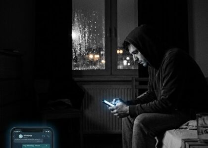 Kapüşonlu bir adam, gece karanlığında yatağın kenarında oturmuş telefon ekranına bakarken; yağmurlu pencerenin ardından şehir ışıkları görünmektedir.