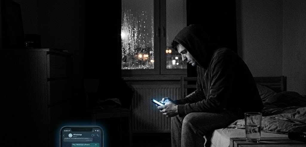 Kapüşonlu bir adam, gece karanlığında yatağın kenarında oturmuş telefon ekranına bakarken; yağmurlu pencerenin ardından şehir ışıkları görünmektedir.